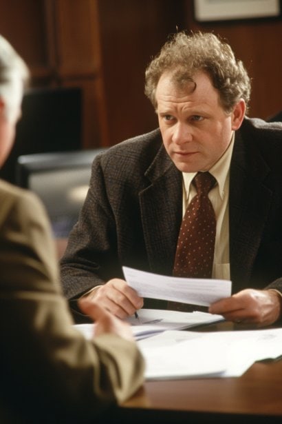 A middle-aged male professional, in a formal suit, having a negotiation meeting with an HR representative. He's calmly but assertively discussing terms, with documents in hand, symbolizing the negotiation process for reemployment.