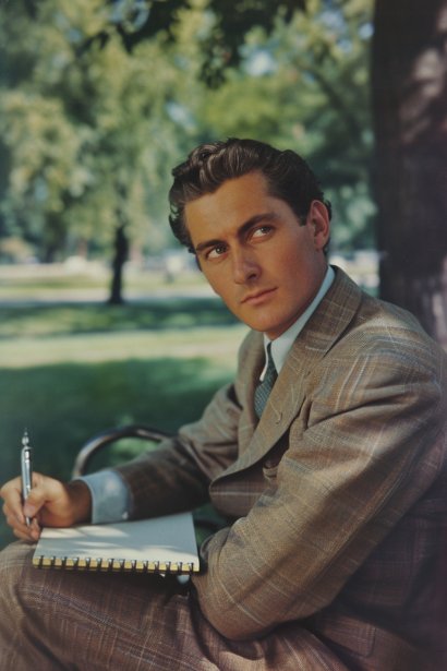 A thoughtful young male professional sitting at a park bench, gazing into the distance, with a notepad and pen in hand. He's jotting down notes, reflecting a moment of introspection and planning for the future after job loss.