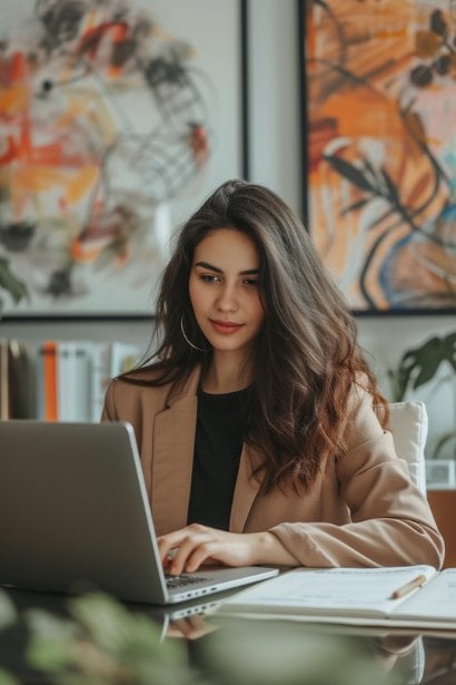 A young female professional, looking reflective and satisfied, sitting at her desk and typing a summary of the meeting on her laptop. She has a notepad beside her with a list of action items and key points, symbolizing the process of documenting feedback and planning next steps.