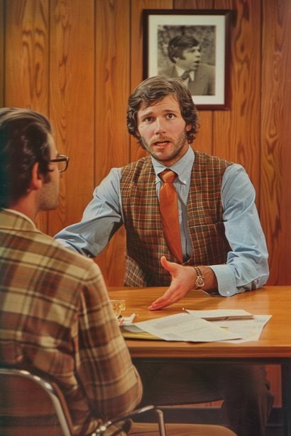 A small meeting room scene with a male employee and his manager discussing across a table. The employee is presenting his case with open hands, showing a clear and respectful communication style. Both individuals are engaged in a serious yet constructive conversation, reflecting a professional negotiation of expectations.
