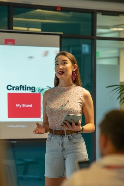 A late 20s female professional, standing confidently at the front of a small, well-lit conference room. She's engaging her audience during a presentation, with a slide behind her titled "Crafting My Brand" Her expression is passionate and persuasive, illustrating the dynamic process of building a personal reputation through effective communication and brand messaging.
