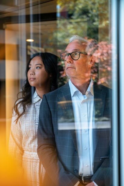 A photo capturing a moment of reflection where a male senior executive and a female junior employee stand by a window, looking out. The atmosphere is contemplative, with the executive displaying an expression of introspection and the employee showing a slight concern. This scene represents the complexities and interpersonal dynamics involved in navigating the power dynamics and biases inherent in reverse shadowing.