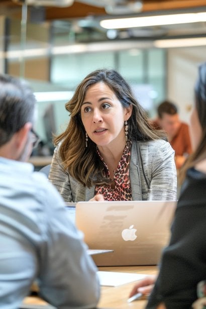 A female professional, appearing resilient and composed, engaged in a deep conversation with her team in a modern, open-space office. The team is gathered around a laptop, discussing strategies with an adaptable approach to a sudden project change.