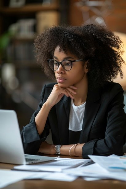 A serious and focused young female entrepreneur sitting at a desk, surrounded by documents and a laptop. She's reviewing papers and appears deep in thought, symbolizing the due diligence and evaluation process in considering a turnkey business.