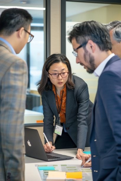 A small, diverse team of professionals, appearing engaged and collaborative, are brainstorming solutions around a laptop in a huddle space. The focus is on a female team leader, looking encouraging and open-minded, as she writes down ideas on a transparent idea board. This image captures the concept of leveraging character strengths to address workplace challenges and drive growth.