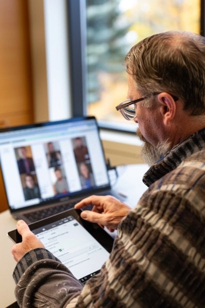 An over-the-shoulder view of a male evaluator reviewing notes on a digital tablet, with the virtual group interview ongoing on a laptop screen in front of him. He appears to be making thoughtful considerations, highlighting the evaluation process in a virtual group setting. Each candidate's frame on the laptop shows them engaged in discussion or awaiting feedback, reflecting the interactive nature of the assessment.