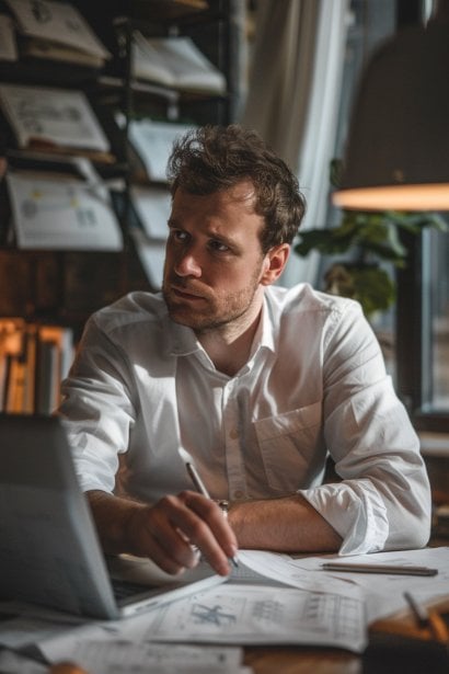 A photo depicting a young male professional, sitting at a desk cluttered with technical drawings and a laptop, deep in thought as he solves a complex problem. His expression is one of intense concentration and determination, illustrating the evaluation of technical and problem-solving skills in a functional interview context.
