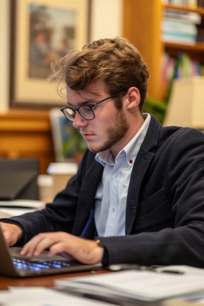 A photo of a young male professional at his desk, typing on a laptop with a focused expression. Papers and notes are spread around him, portraying him in the act of following up on meeting decisions and actions, ensuring continuity and impact.