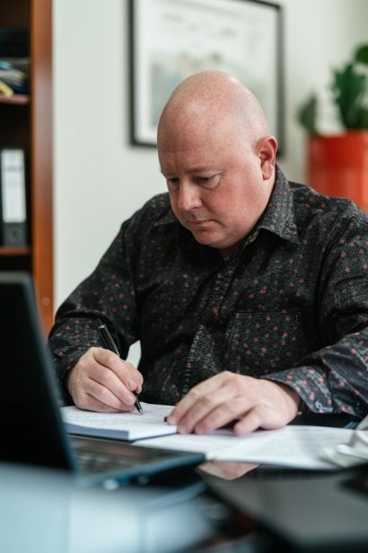 A photo of a male professional at his desk, writing a note before sending a text. His expression is concentrated and respectful, highlighting the meticulousness required in crafting a polite cancellation message. The office setting is tidy and well-lit, reflecting a professional environment.