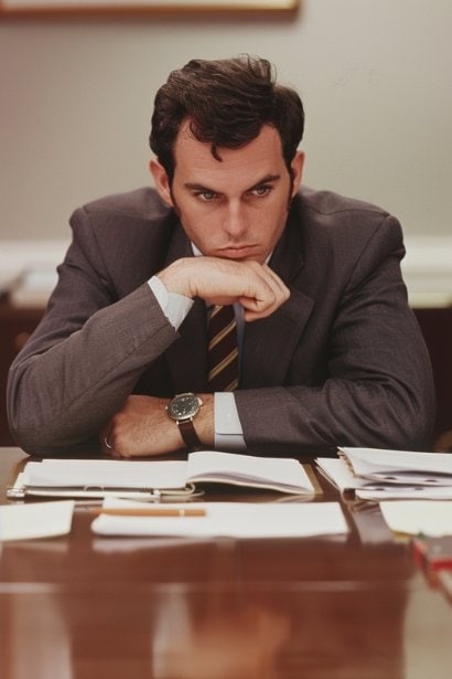 A middle-aged male professional sitting at a conference table, looking slightly anxious with his hands clasped together. His eyes are focused on some papers in front of him, symbolizing his anticipation and the common fear of speaking in meetings.