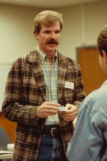 A middle-aged male professional, appearing proactive and engaged, in a seminar room, exchanging business cards with another professional. The background shows a seminar in progress, emphasizing ongoing professional development and networking during the returnship.